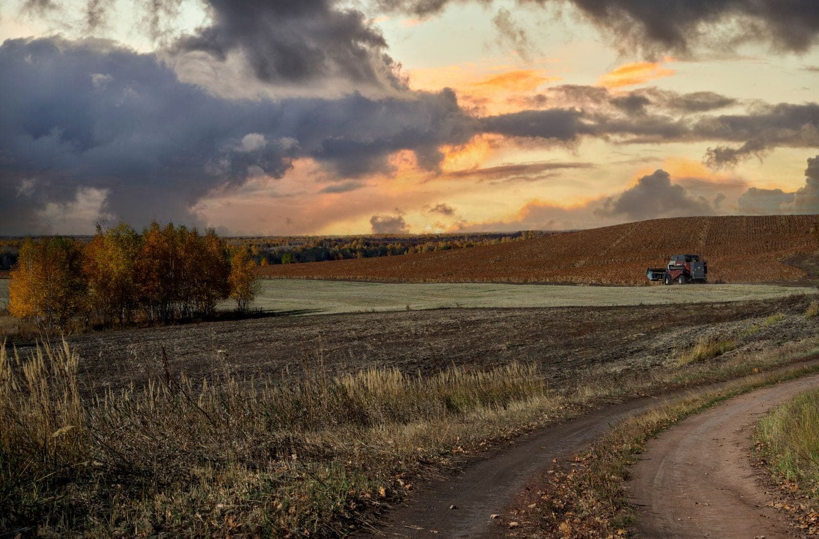 A tractor navigating vast autumn croplands against a dramatic sunset sky