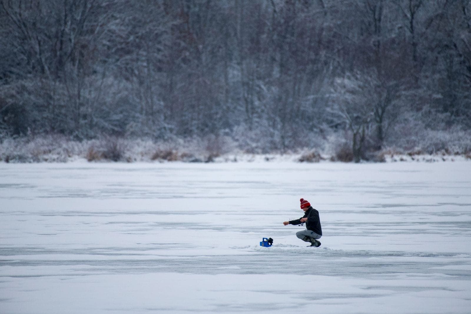 A lone fisherman ice fishing on a frozen lake surrounded by snow-covered forest.